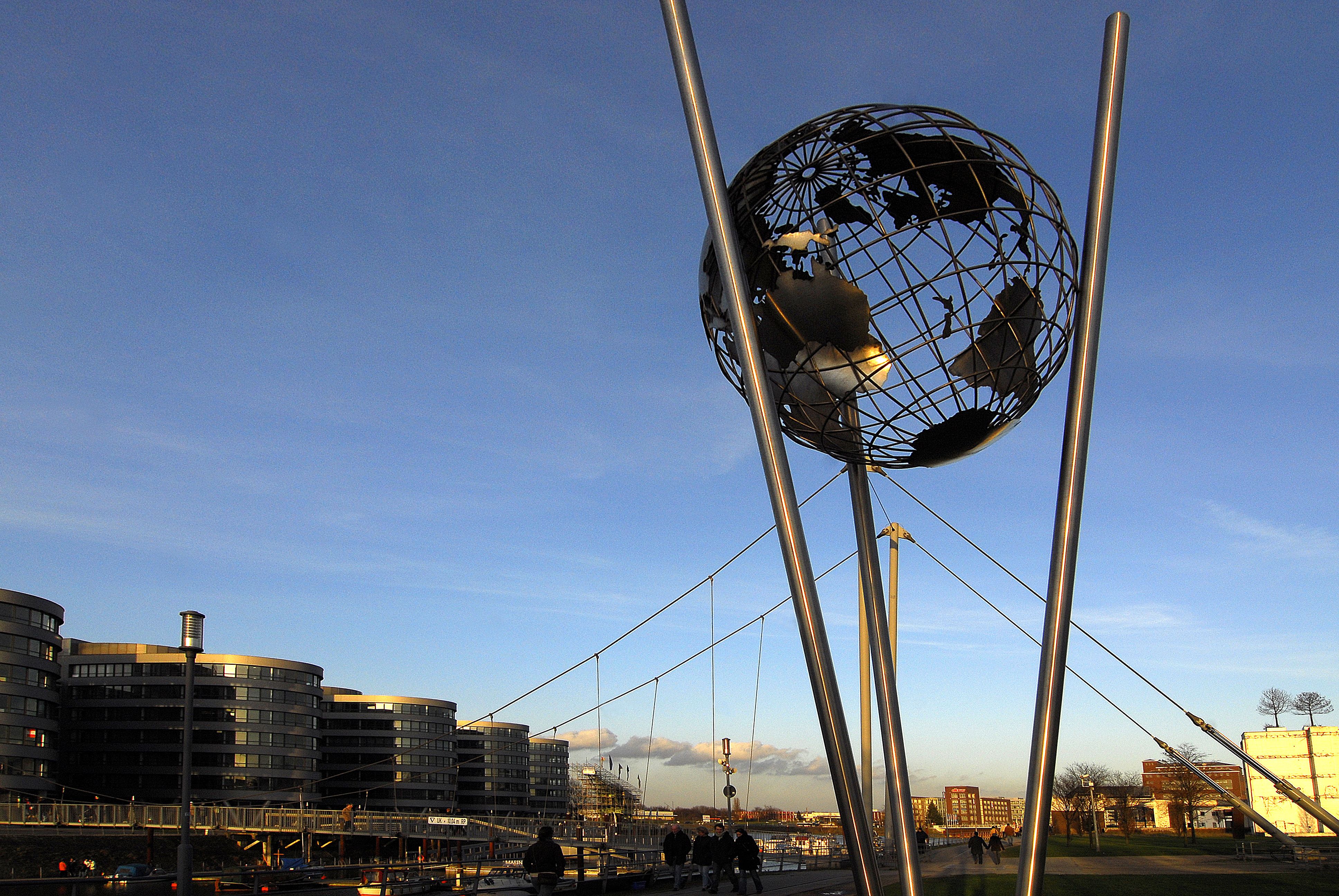 Skulptur eines Metallglobus vor blauem Himmel und Hafengebäuden