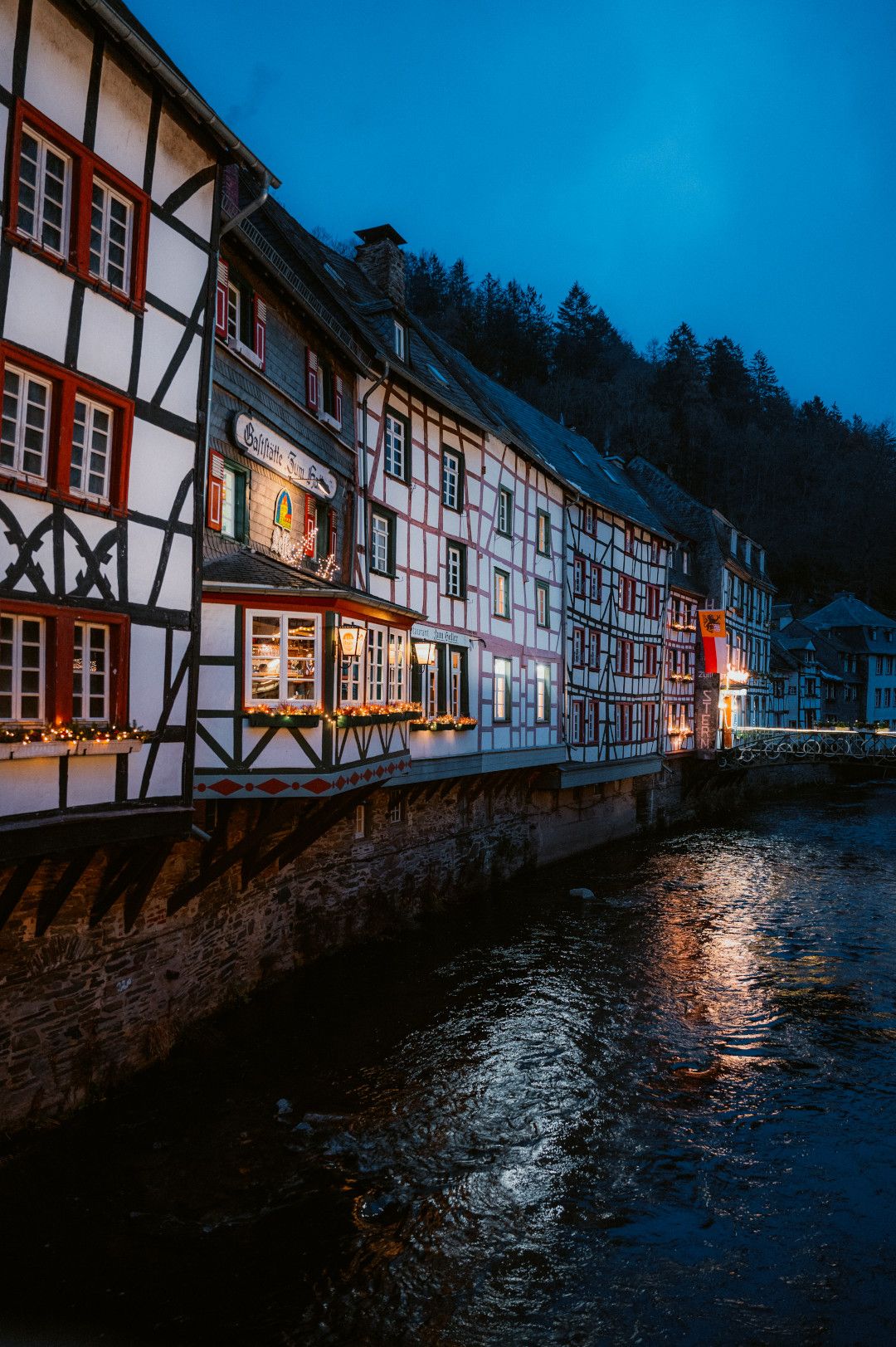 Beleuchtete Fenster von Fachwerkhäusern spiegeln sich im Wasser des vorbeifließenden Flusses wieder